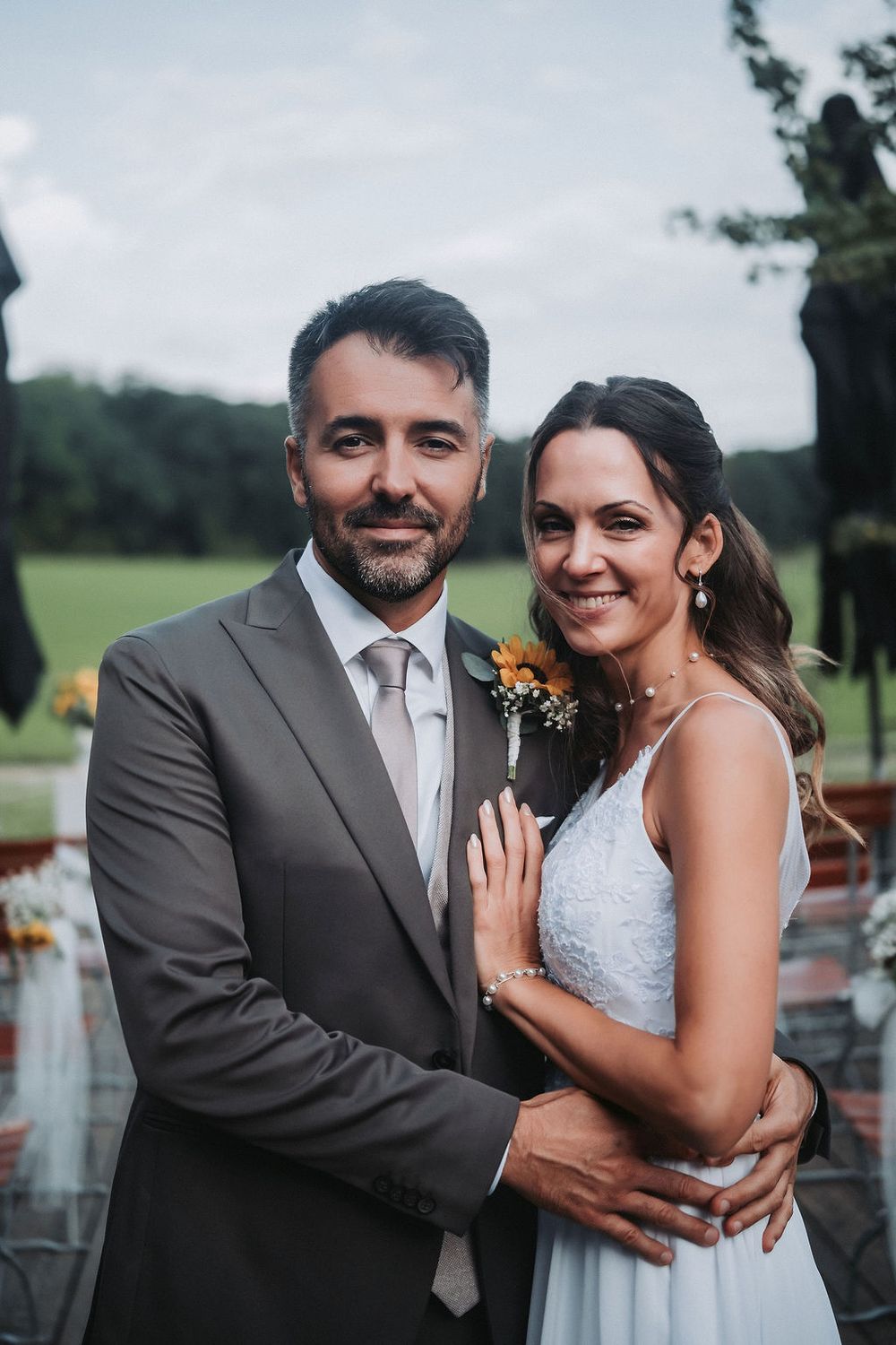 A couple in wedding attire embrace outdoors, with the bride in white and groom in gray suit with orange boutonniere.