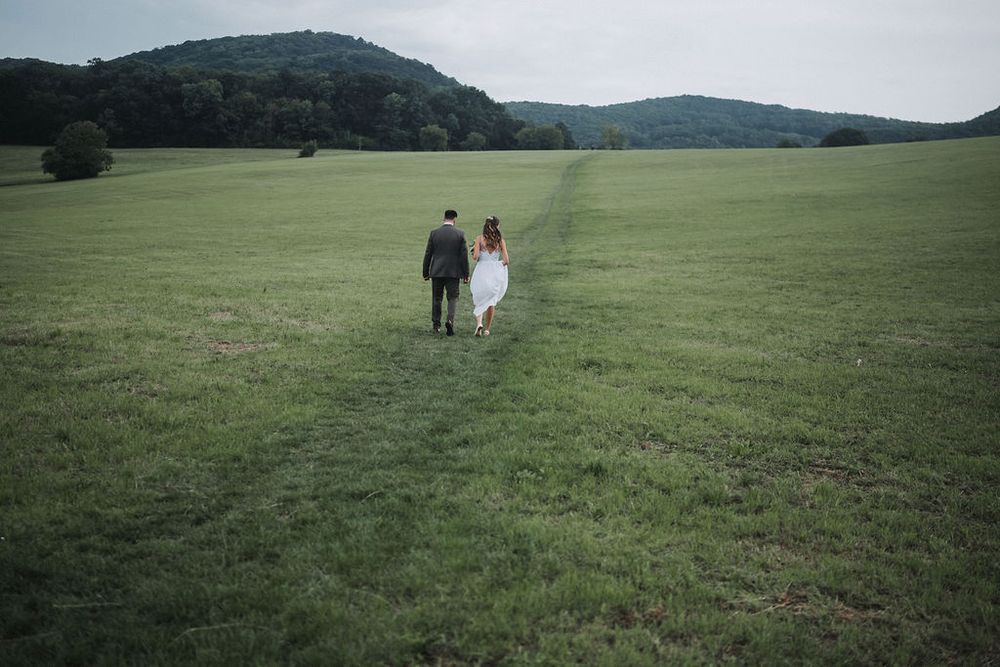 Two figures walk hand in hand across a vast green meadow with rolling hills in the background on a cloudy day.