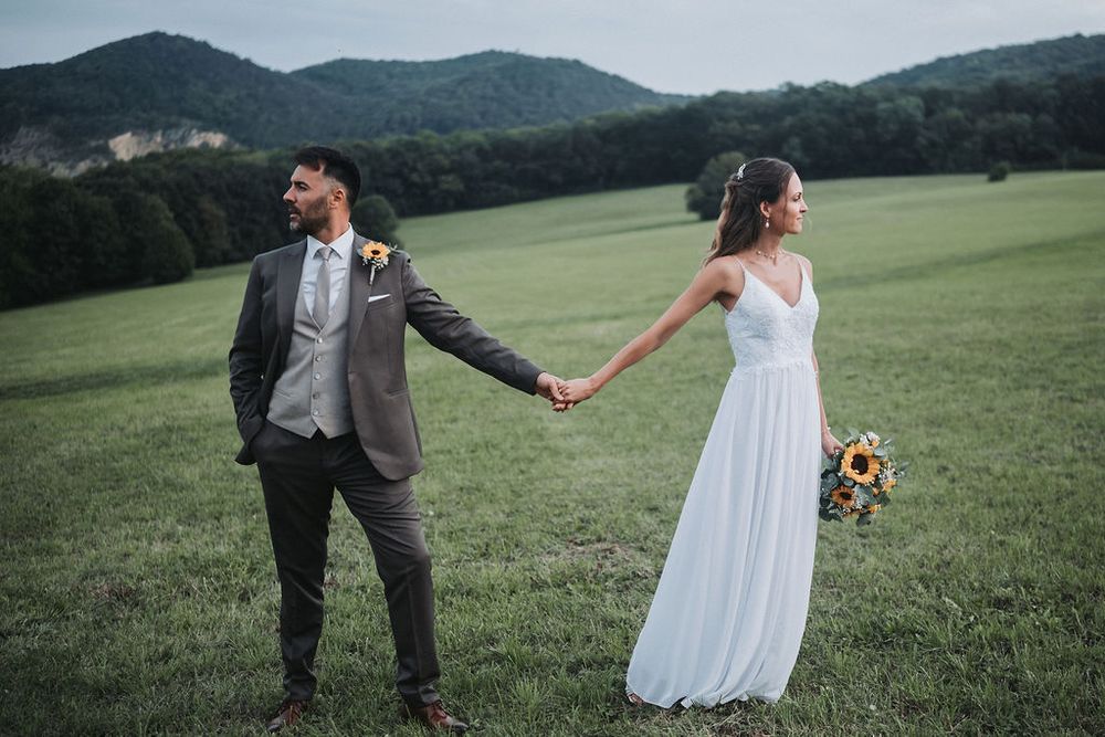 A couple in wedding attire holds hands while walking through a lush green meadow with mountains in the background.
