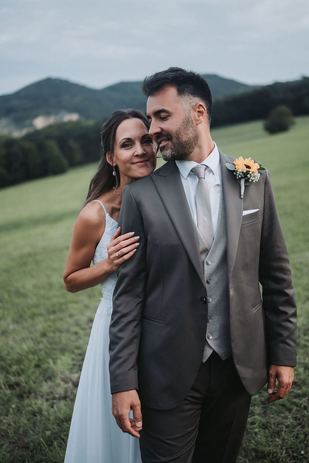 Romantic wedding couple embraces in a scenic mountain meadow wearing elegant gray suit and white dress.