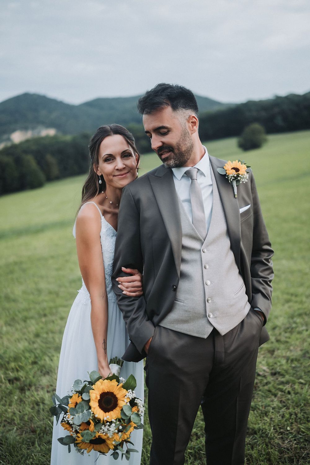 Couple shares intimate moments in a green field with sunflower bouquet during their outdoor wedding photoshoot.