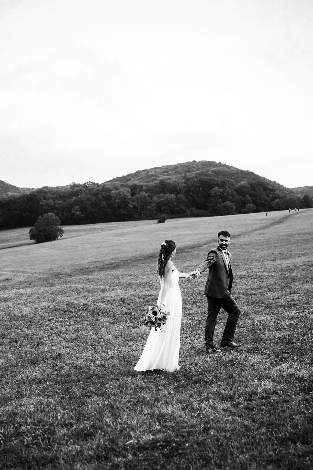 A couple in wedding attire playfully runs through a grassy meadow with rolling hills in the background.
