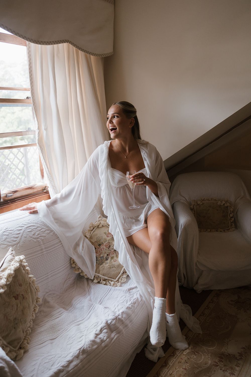 A person in a white dress sits by a window on a cozy white couch with decorative pillows in a bright, airy room.