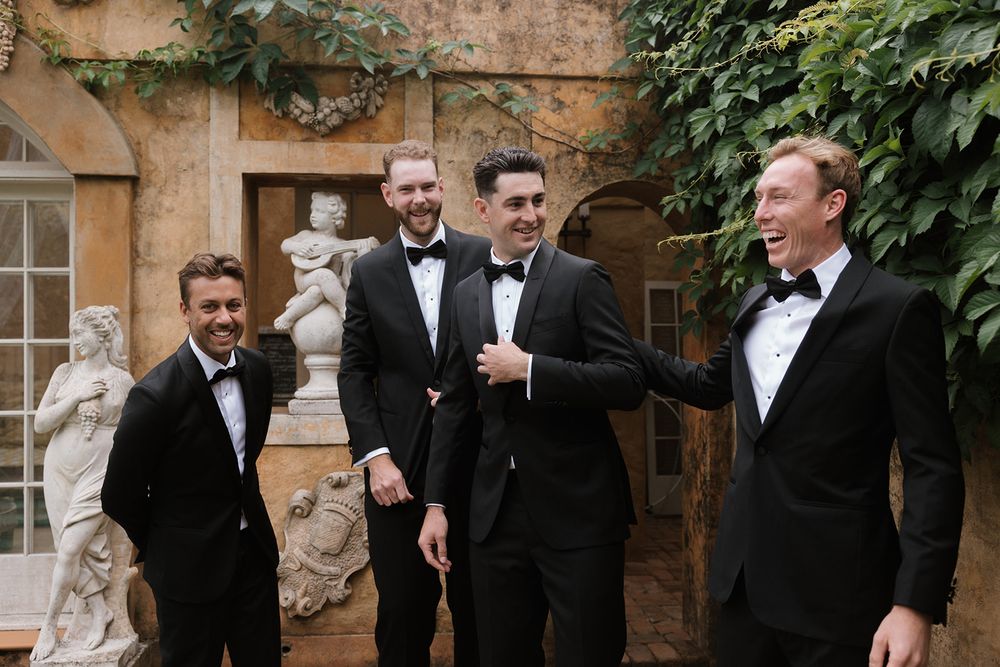 A wedding party in black tuxedos shares a candid laughing moment together outside a rustic stone building.