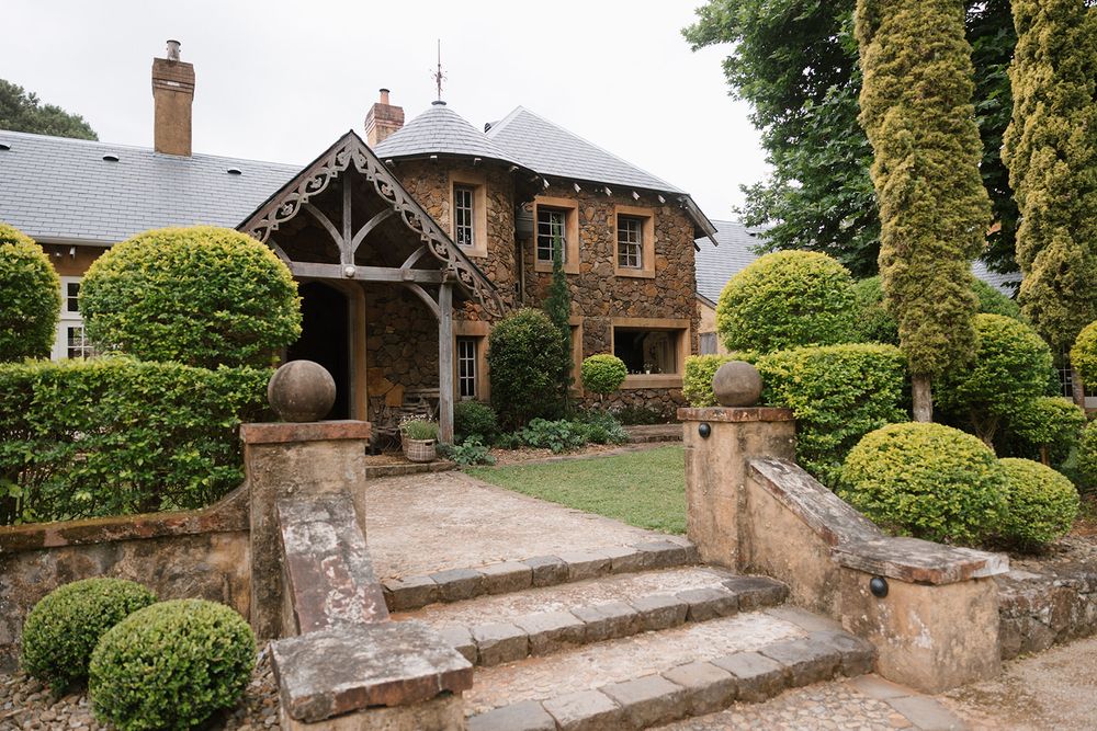 Charming stone cottage with wooden portico entrance and manicured topiary garden featuring stone steps and pillars.