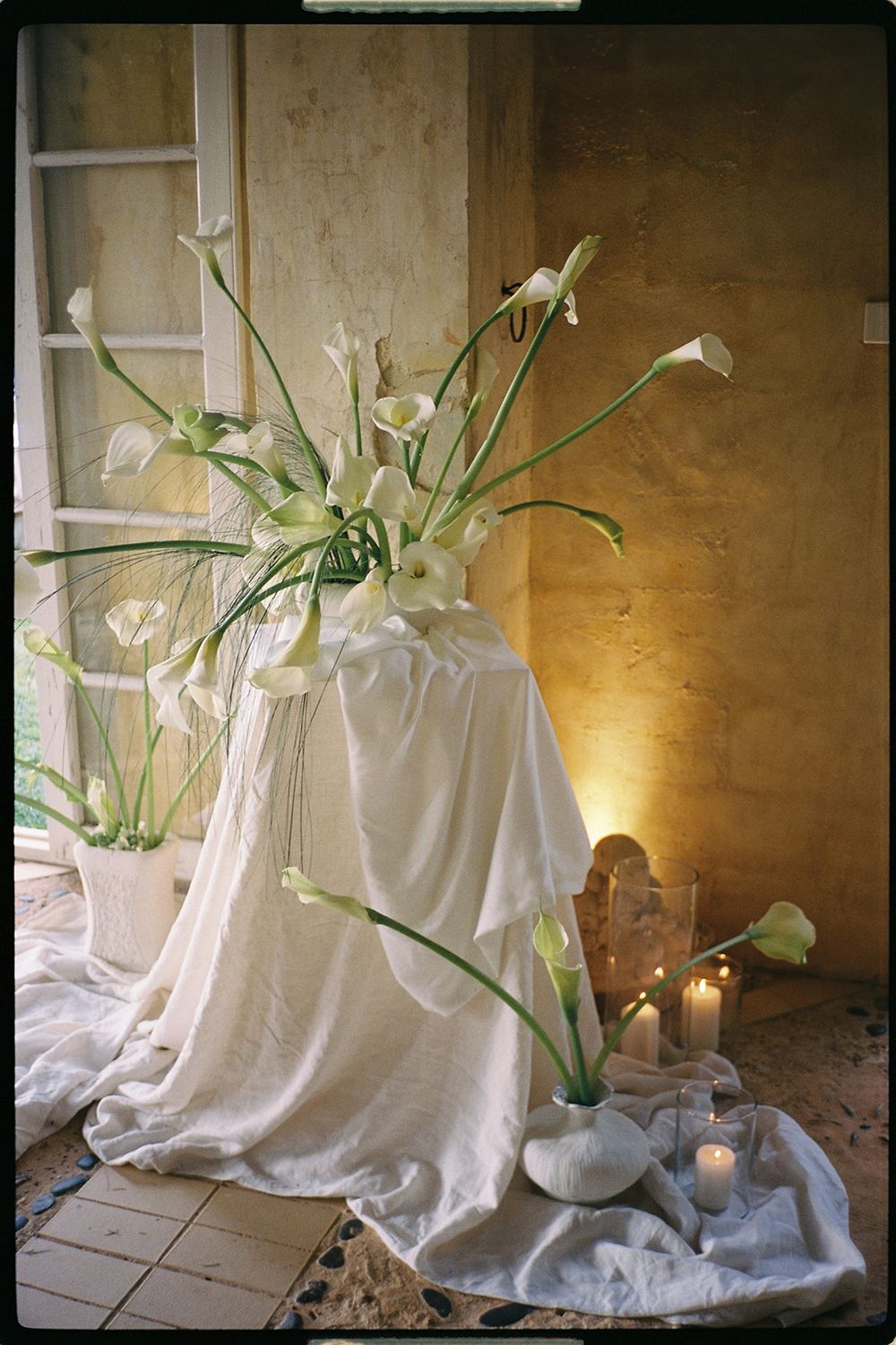 White calla lilies in a draped vase arrangement with glowing candles against a warm textured wall.