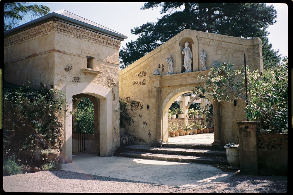 Historic stone gateway with arched entrance, religious statue, and lush greenery at an old European estate.