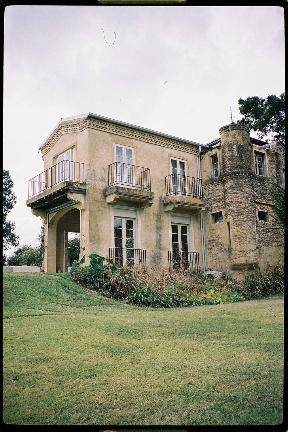 Historic two-story stone mansion with balconies, arched portico, and turret surrounded by green lawn.