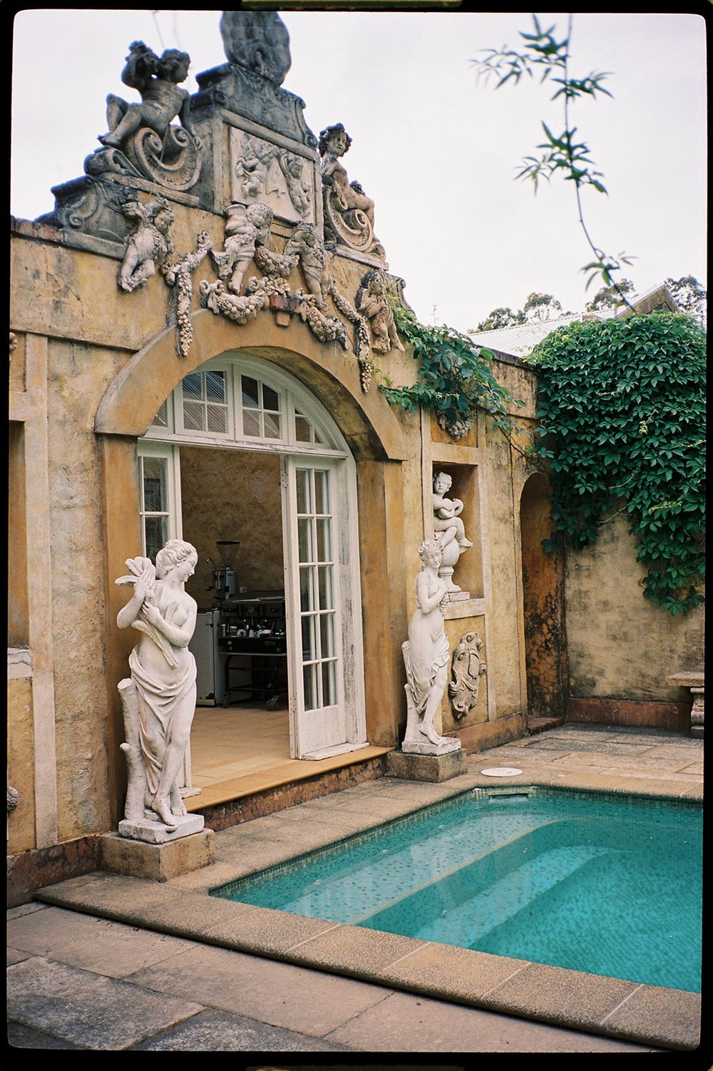 Elegant classical stone statues flank a ornate baroque archway beside a turquoise pool with ivy-covered walls.