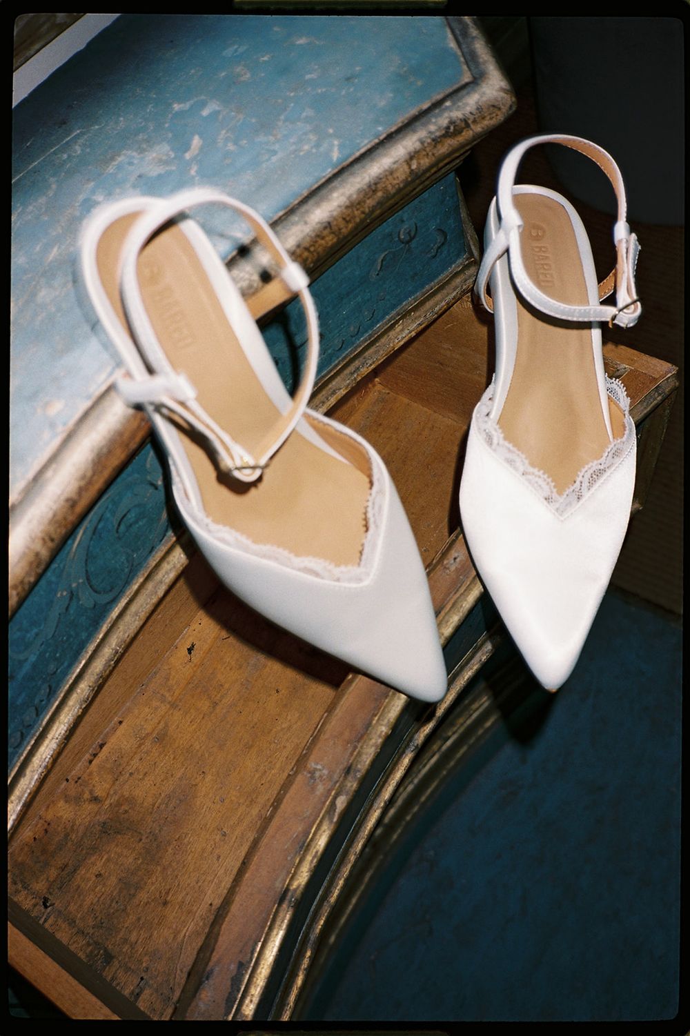 White satin pointed-toe kitten heel wedding shoes with delicate lace trim and ankle strap, displayed on vintage wooden furniture.