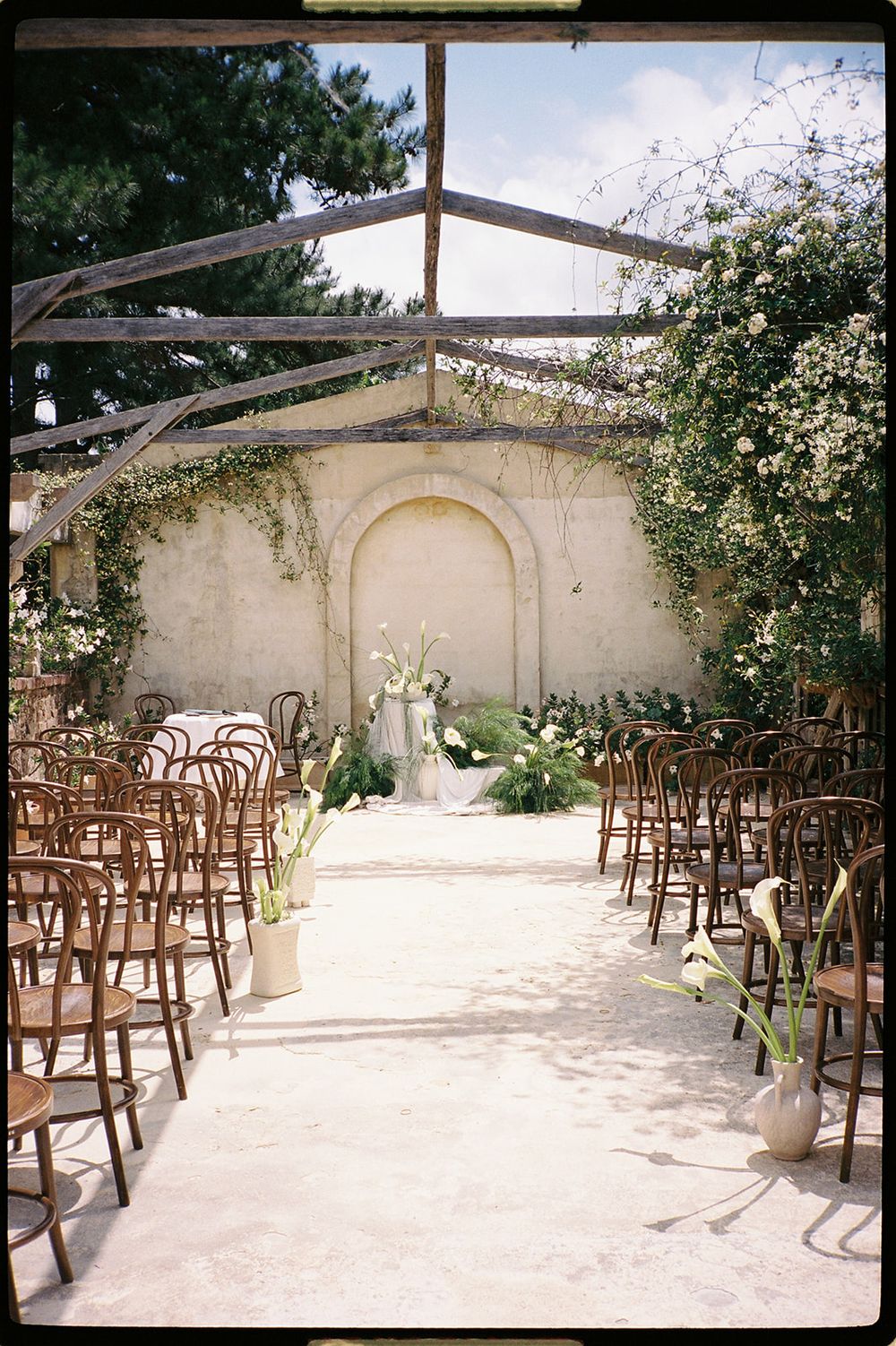 Rustic outdoor wedding ceremony setup with wooden chairs, floral arrangements, and ivy-covered stone wall altar in a sunlit courtyard.