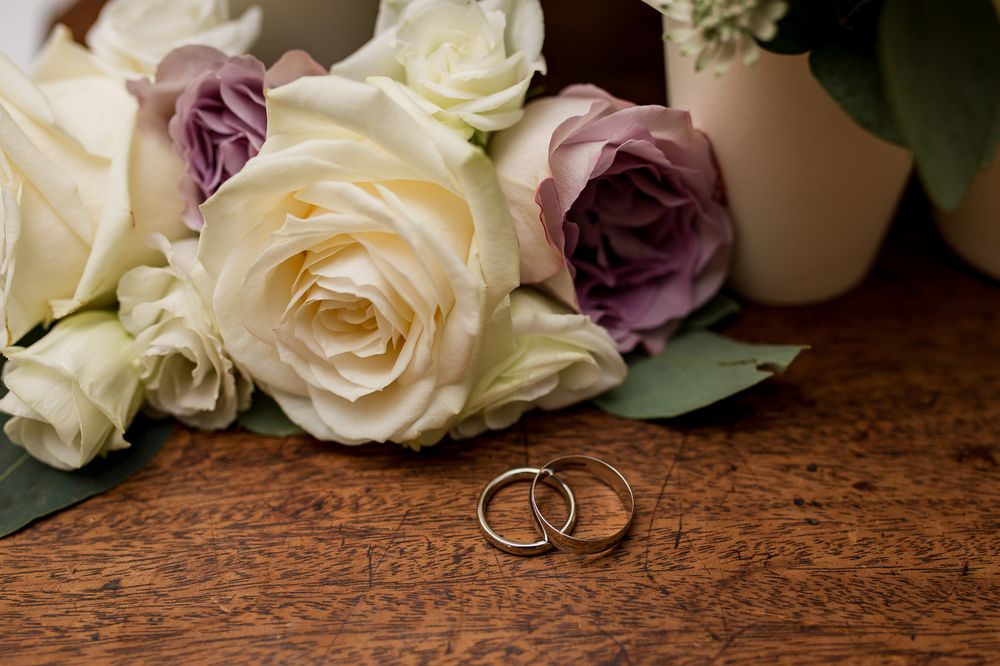 Wedding rings displayed with white and purple roses on a wooden table.