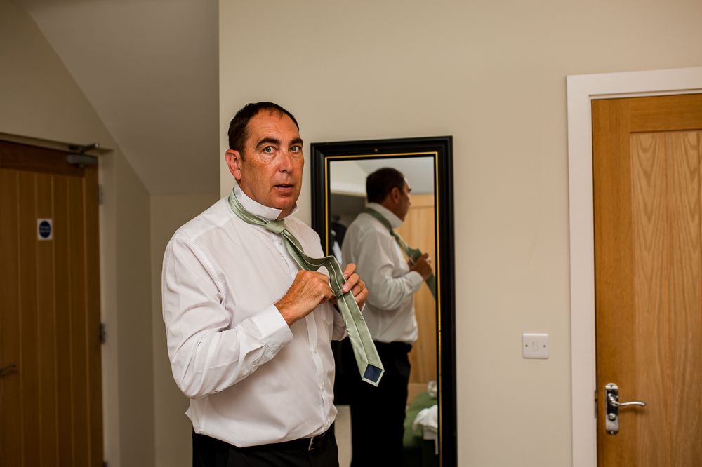 Two men in white dress shirts helping each other with formal attire preparations in a room.