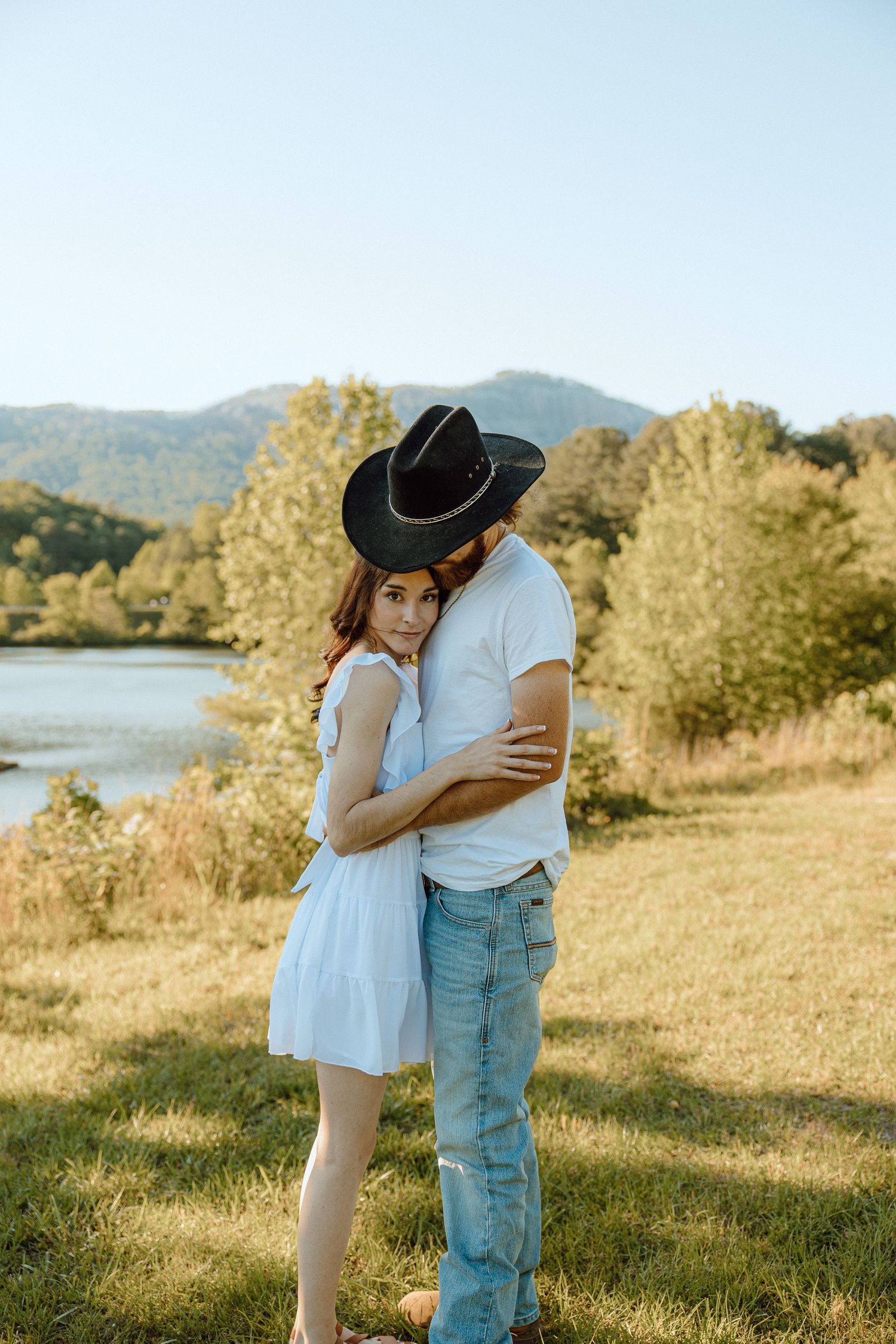 Couple embracing in casual attire on a grassy hillside overlooking a lake.