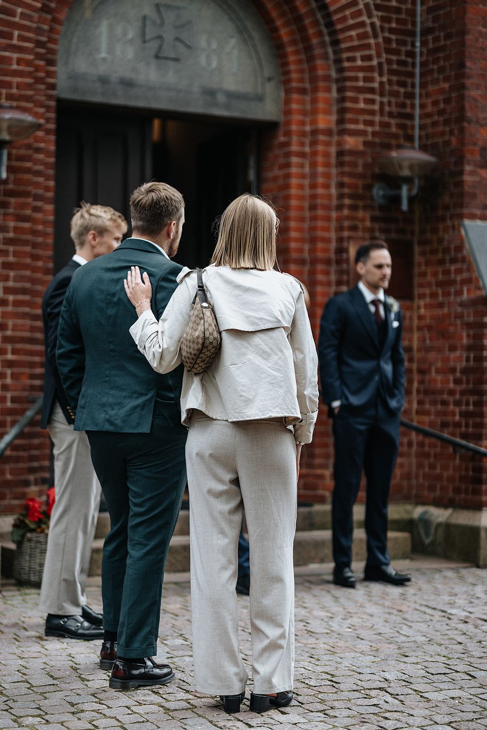 Wedding guests in formal attire gather on a brick courtyard outside a historic building.