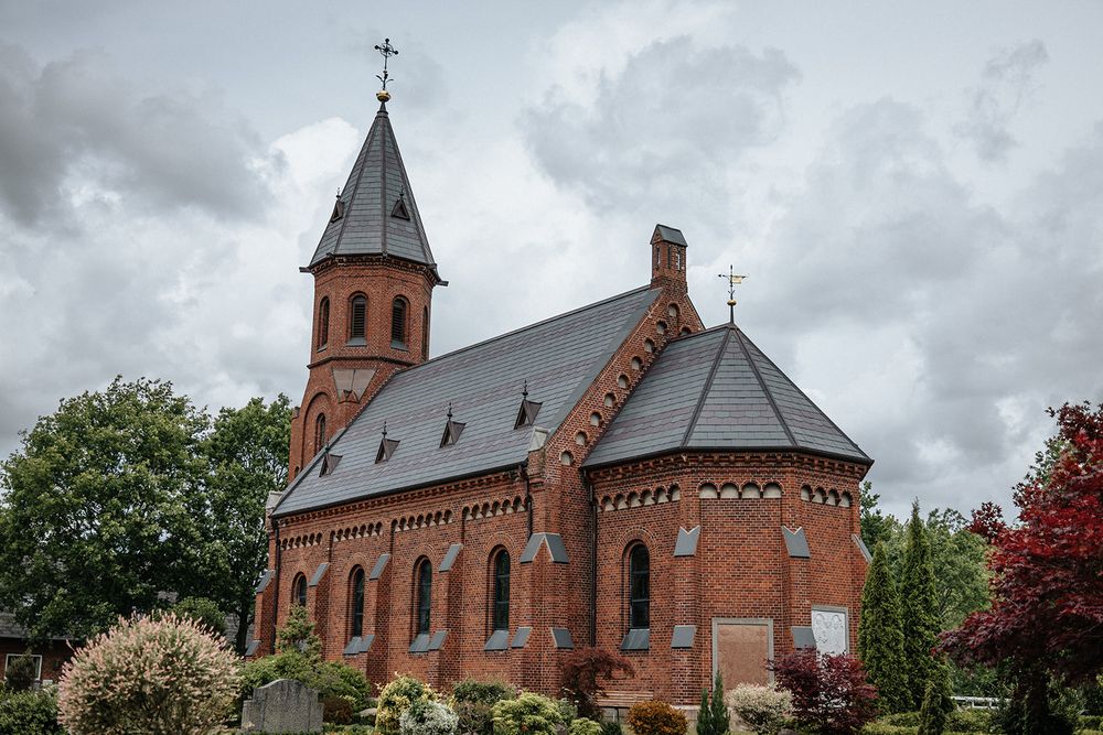 Historic red brick Gothic church with tall spire and ornate architectural details stands under dramatic cloudy sky.