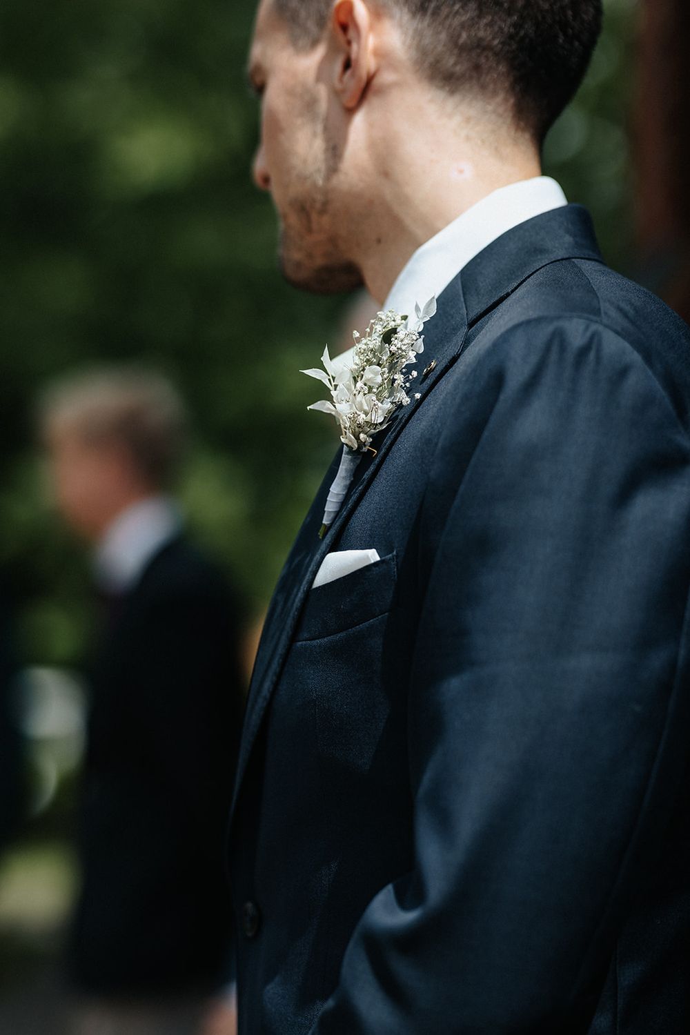 Row of groomsmen in dark suits with white boutonnieres stand in an outdoor garden setting.
