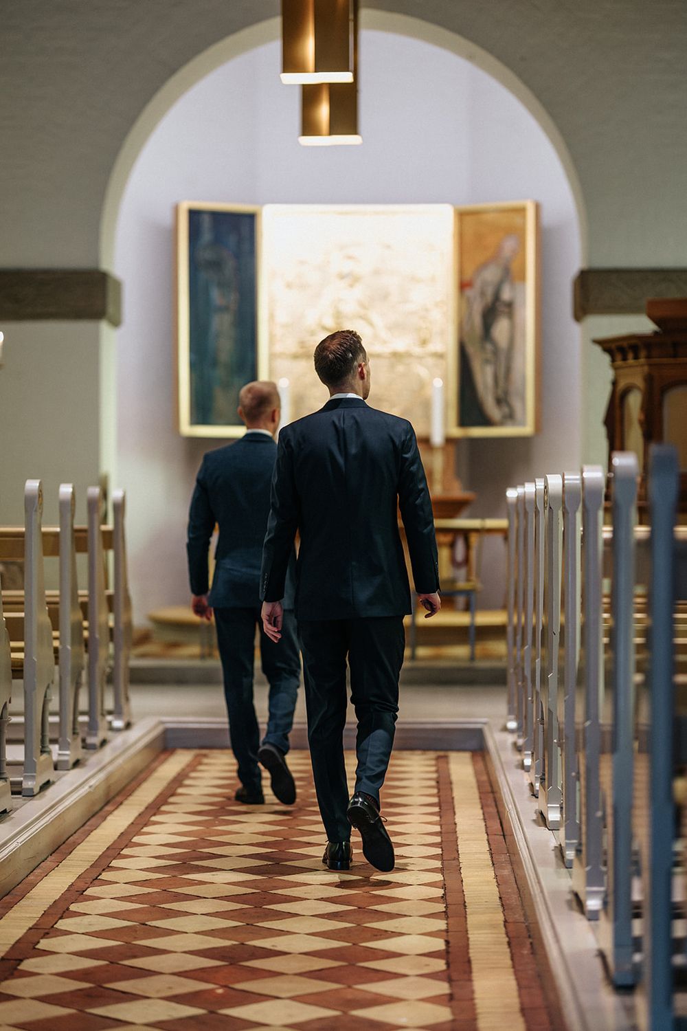 Two people in dark suits walk down a long corridor with arched ceilings and patterned tile floors.