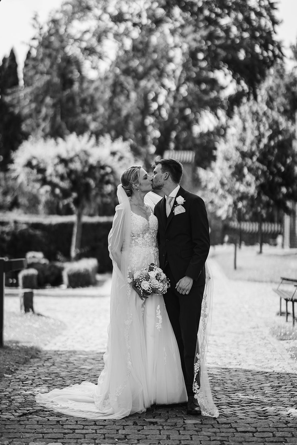 Black and white wedding photos of newlyweds sharing romantic moments on a cobblestone path in an outdoor garden setting.