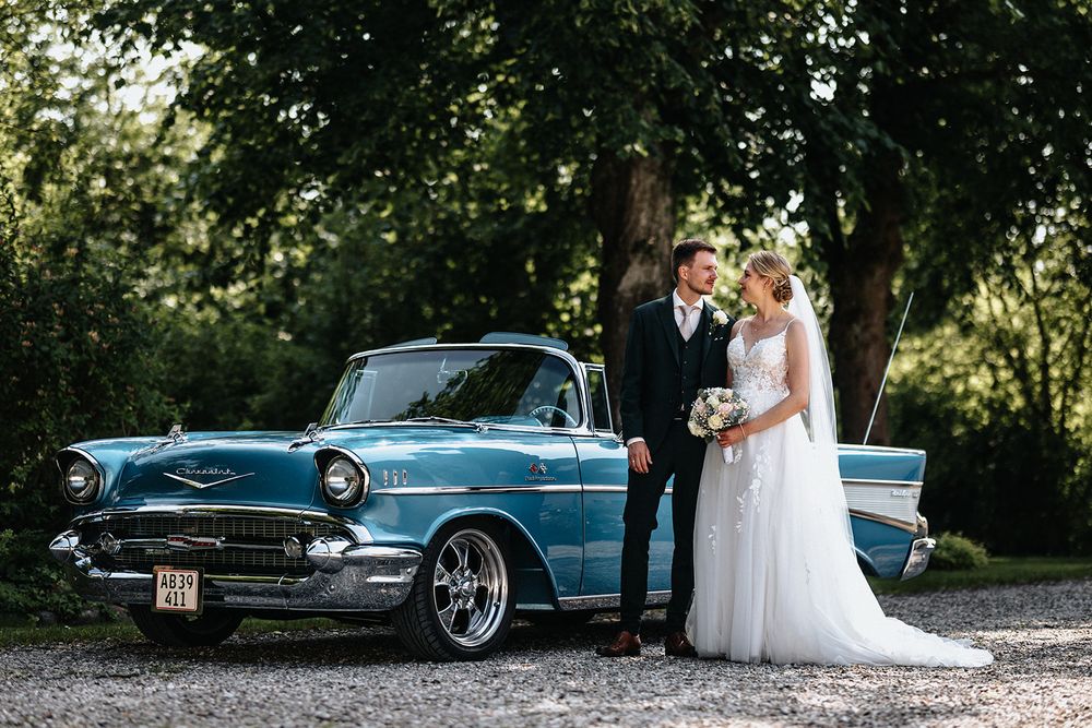 A classic blue vintage car serves as a backdrop for a wedding couple in an outdoor garden setting.