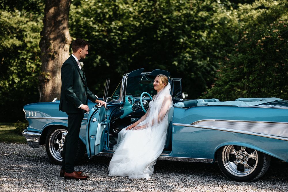Bride and groom pose elegantly beside a classic blue vintage car during their wedding photoshoot.