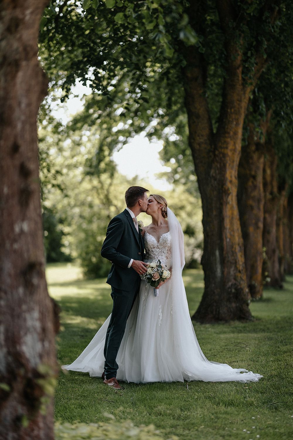 A romantic wedding couple shares an intimate moment under tall trees in a serene outdoor garden setting.