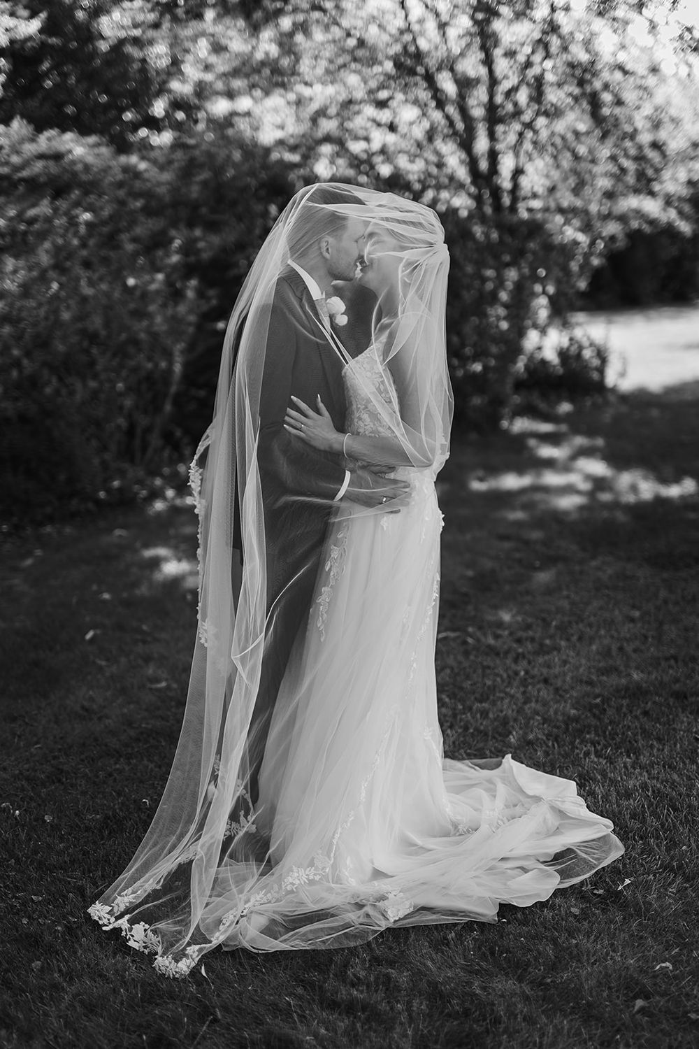 A romantic black and white wedding photo showing a couple embracing under a flowing veil in an outdoor garden setting.