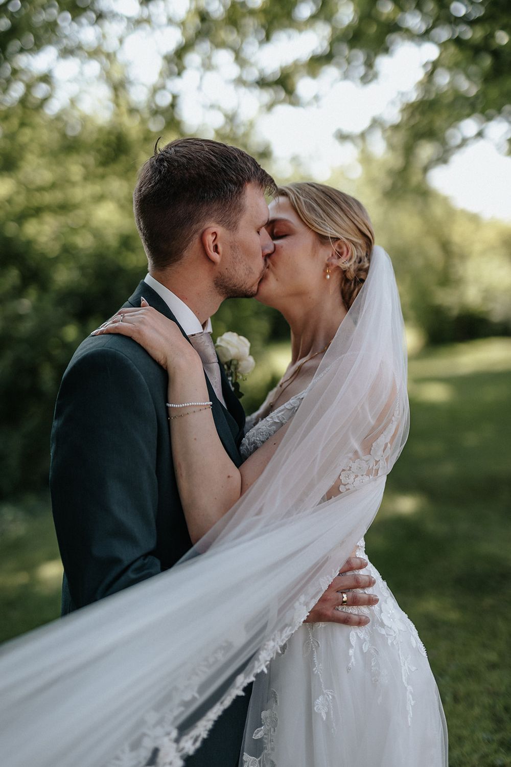 A romantic wedding couple shares a tender kiss outdoors while the bride's veil flows gracefully in the breeze.