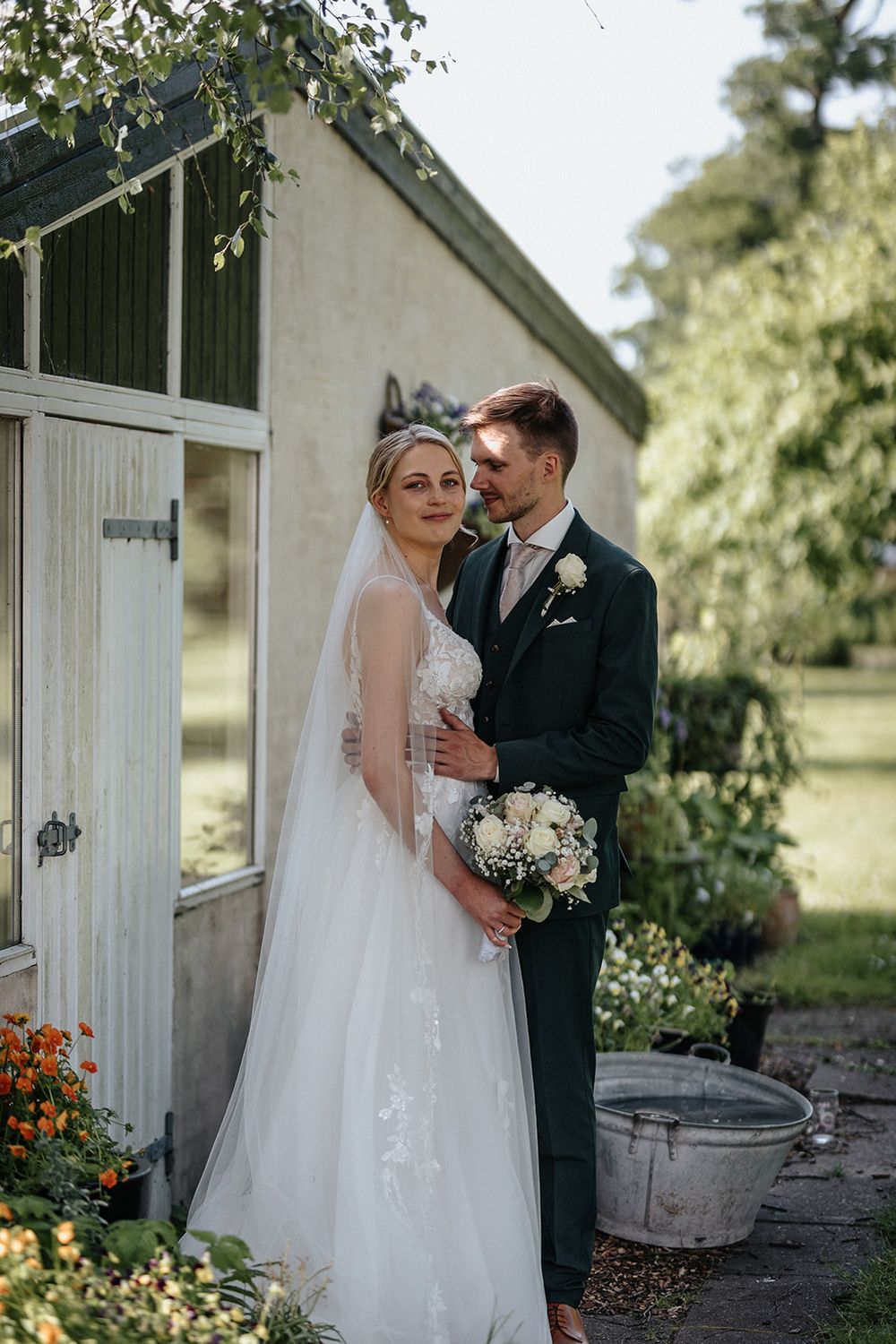 A couple in wedding attire poses together outside a rustic white barn with flowering gardens on a sunny day.