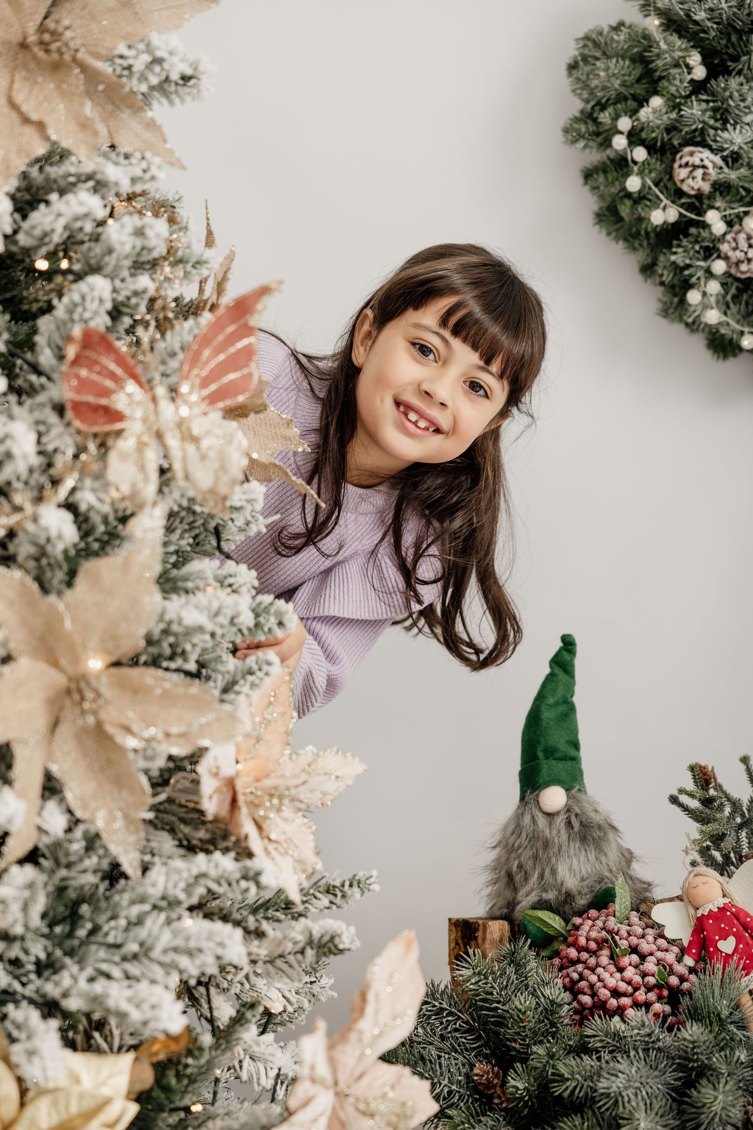 A child sits on a white floor playing with a ukulele next to a decorated Christmas tree with ornaments and decorations.