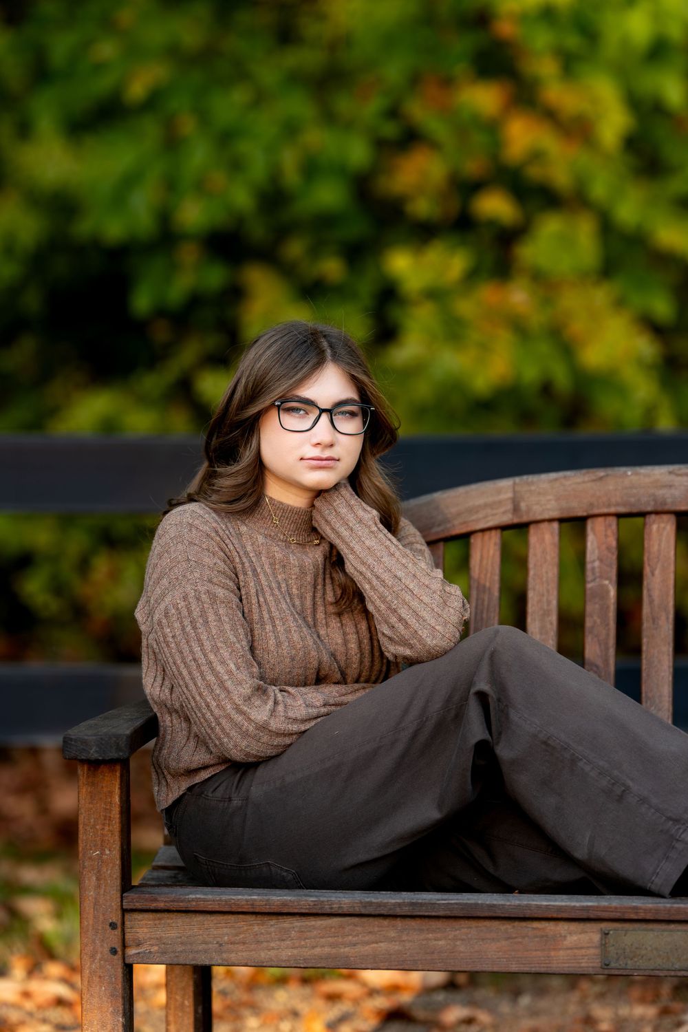 Thoughtful teen girl in glasses and brown sweater sits on wooden bench with vibrant fall backdrop.