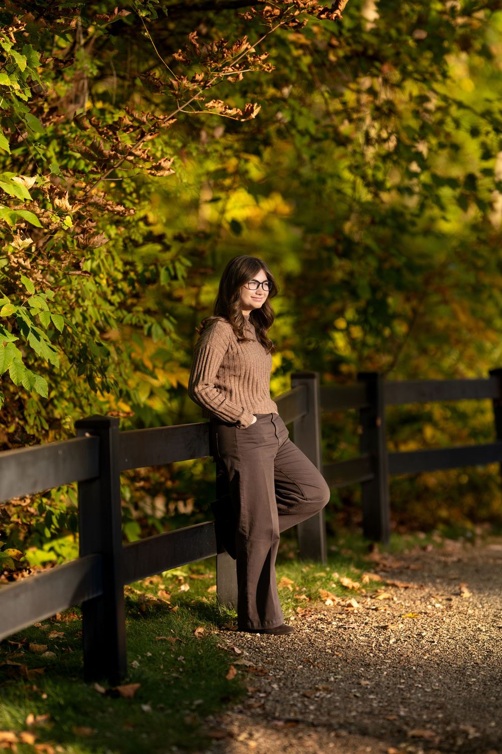 Teen girl in glasses poses by black fence on sun-dappled path with vibrant autumn trees behind.