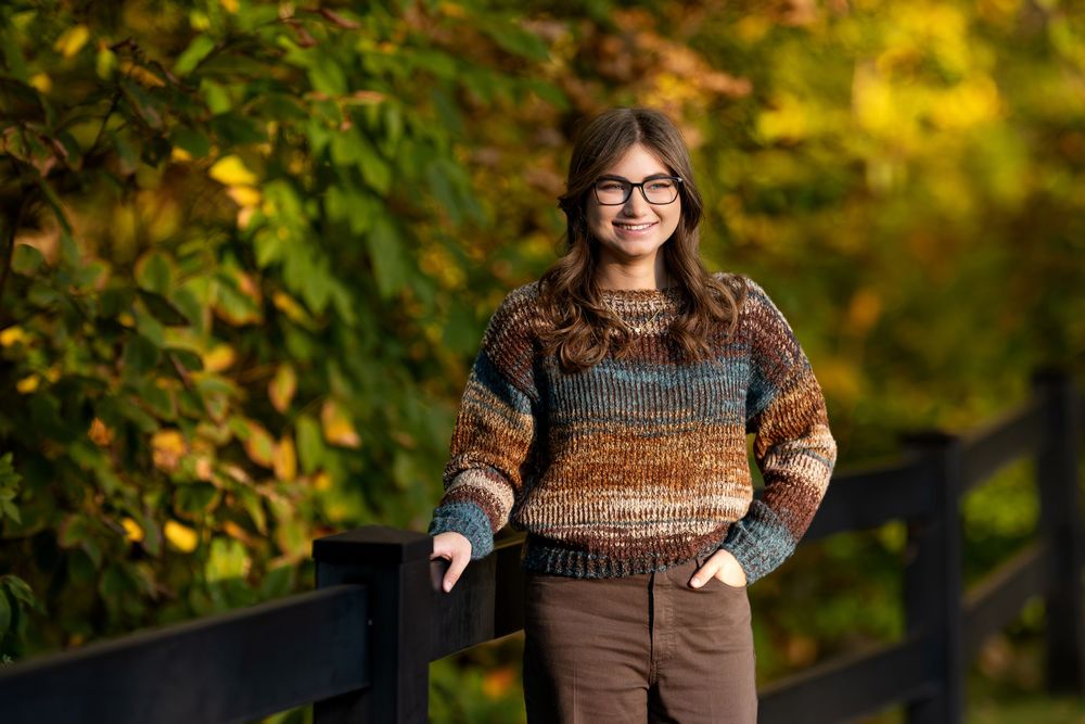 Smiling teen girl wearing colorful striped sweater and glasses poses by black fence with fall leaves.