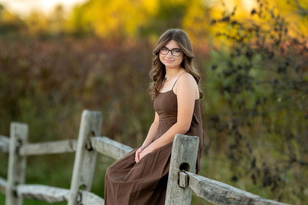 Teen girl with glasses in brown dress sitting on weathered white fence with fall foliage background.