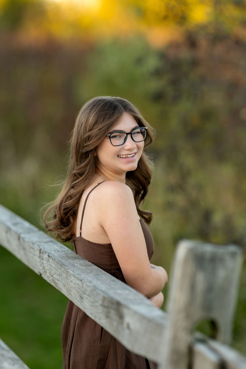 Laughing young woman with glasses in brown dress leaning on wooden fence at golden hour.
