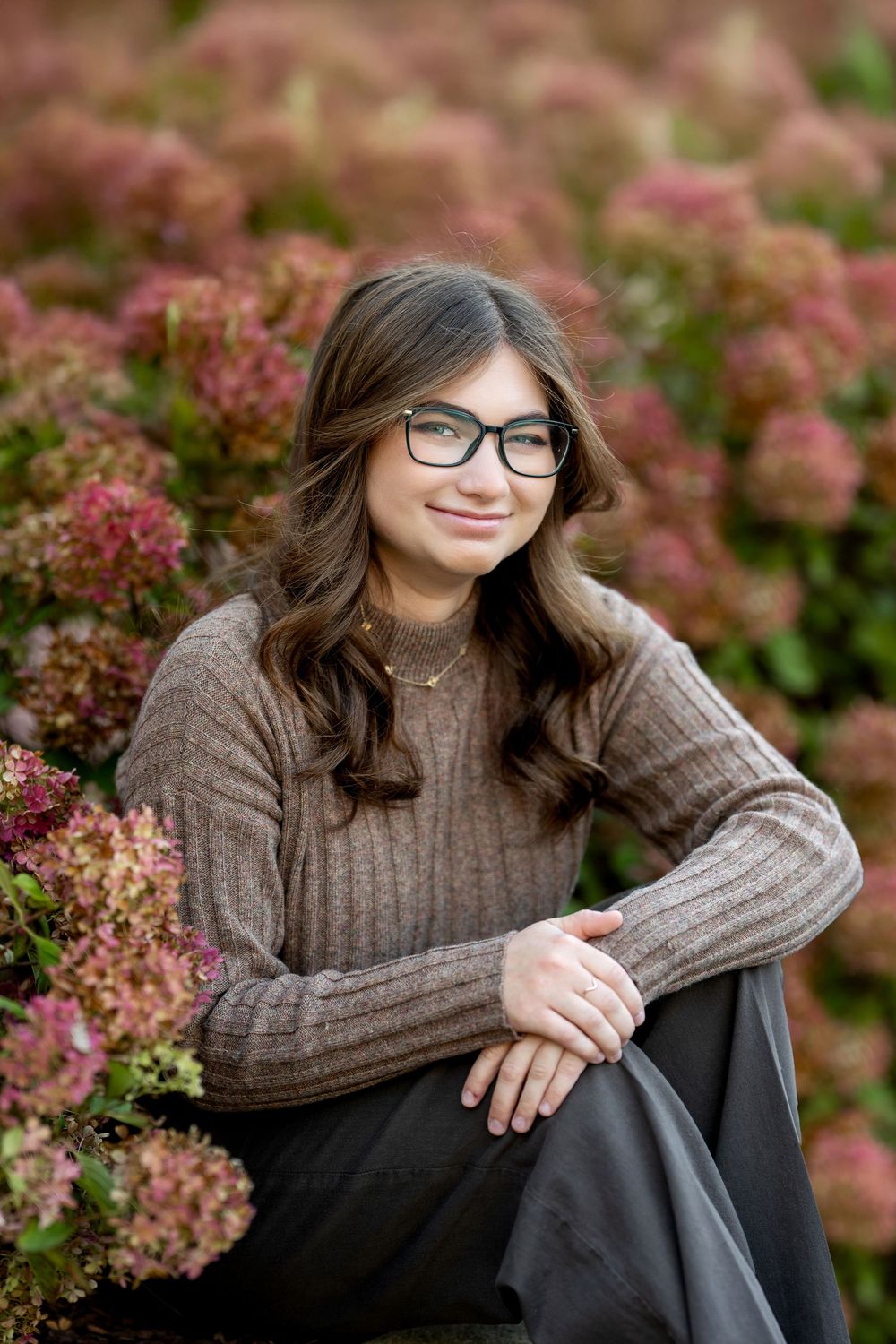Teen girl in brown sweater and glasses sits among pink hydrangeas in a fall garden portrait.