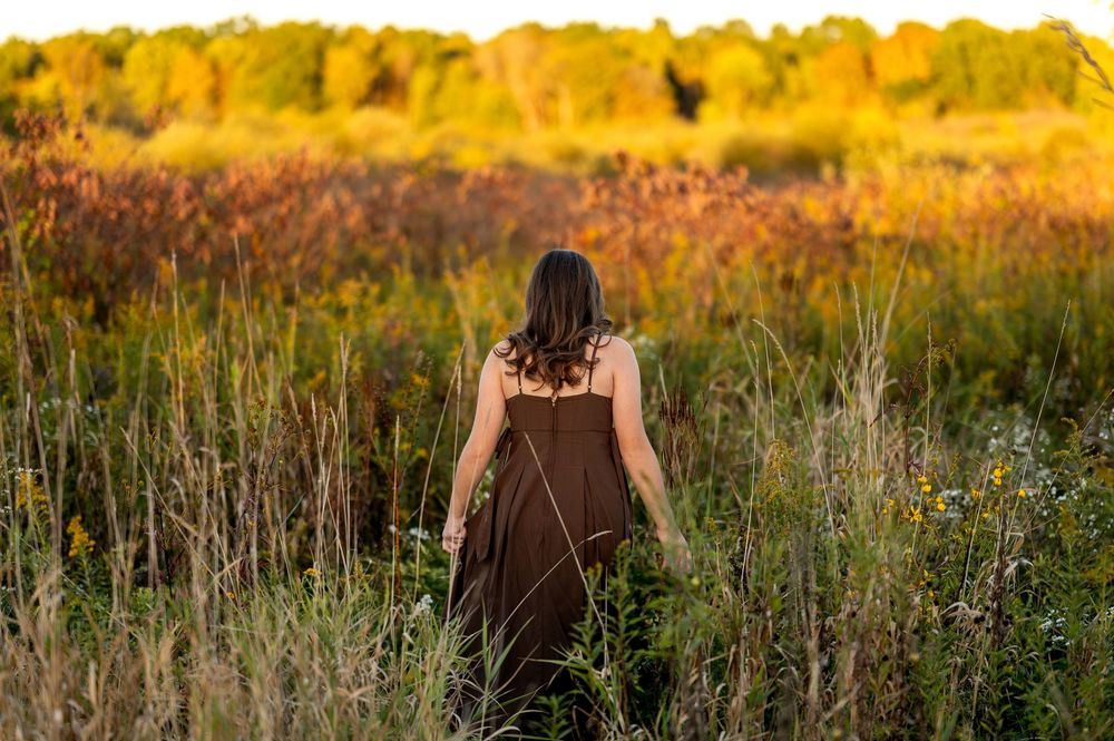 Back view of young woman in brown dress walking through golden autumn meadow at sunset.