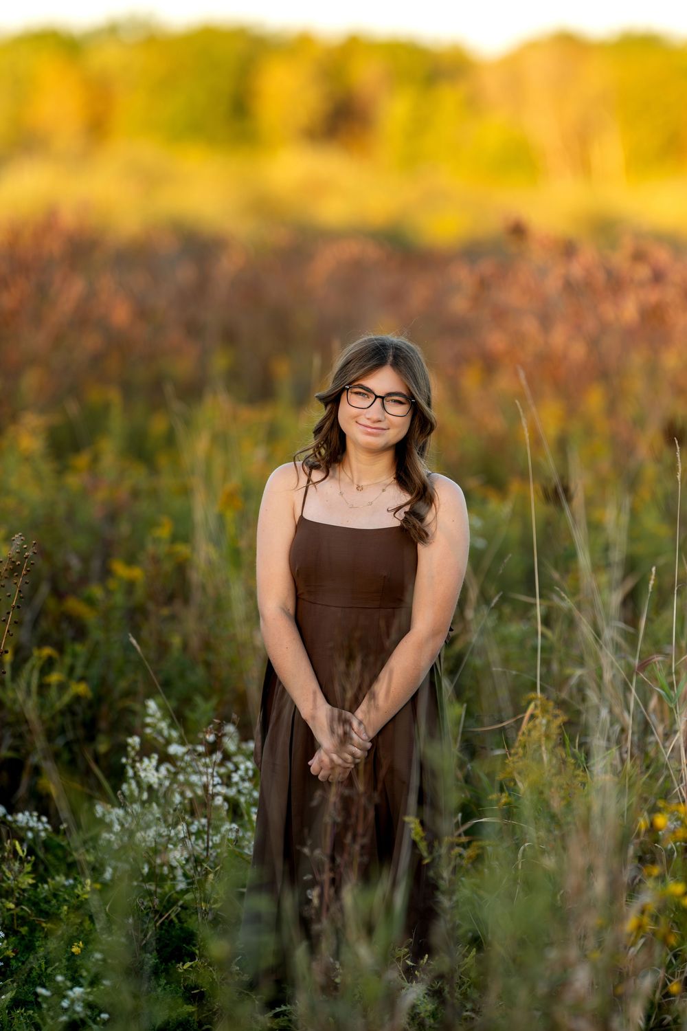 Smiling teen girl in brown dress and glasses standing in golden wildflower meadow at sunset.