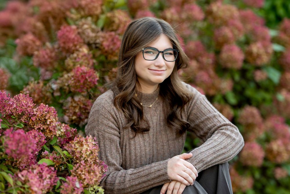 Young woman with glasses smiles beside colorful autumn hydrangea bushes in an outdoor photo session.