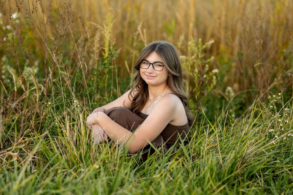 Smiling teen girl with glasses sitting in tall golden grass wearing a brown dress.