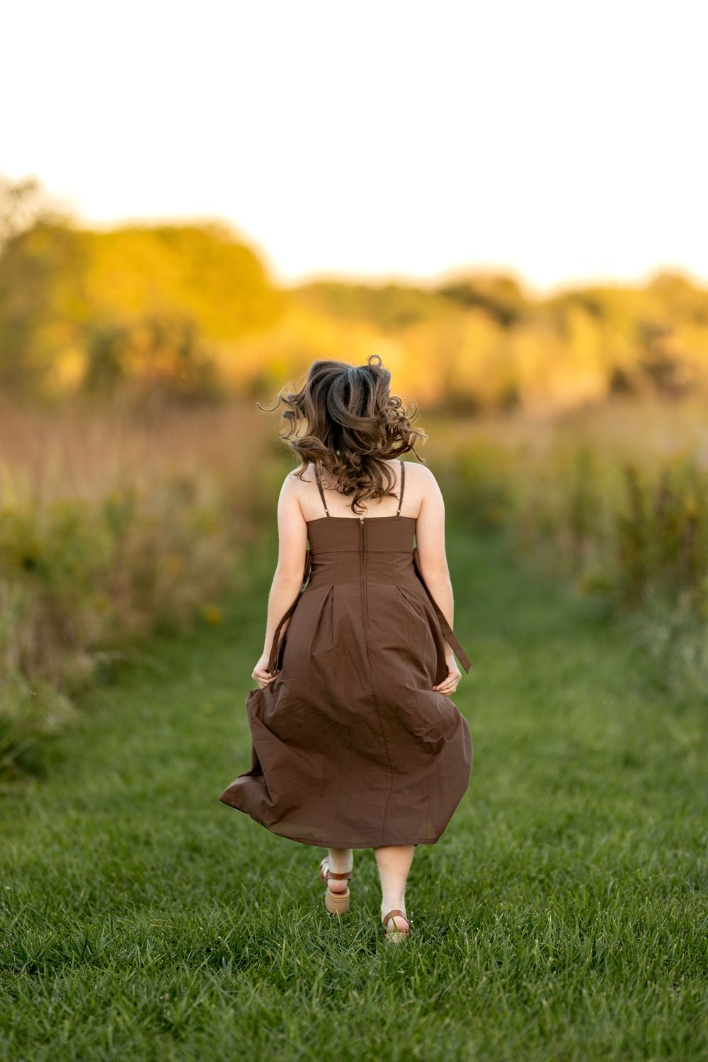 Young woman in brown sundress walking away through a green meadow path at sunset.