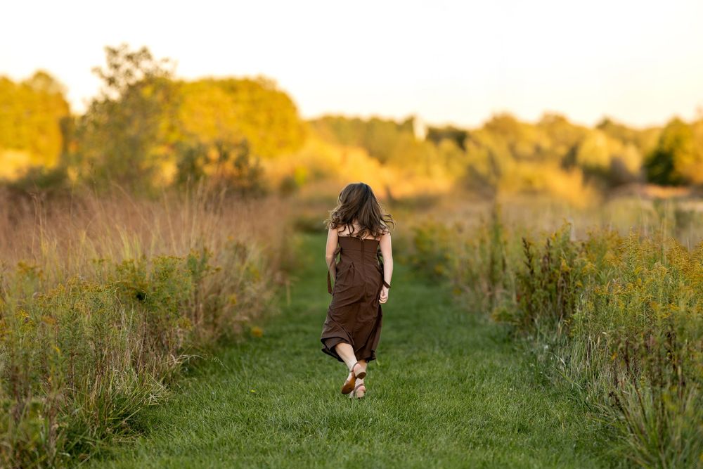 Teen girl in brown dress walking down a grassy path surrounded by golden sunset fields.