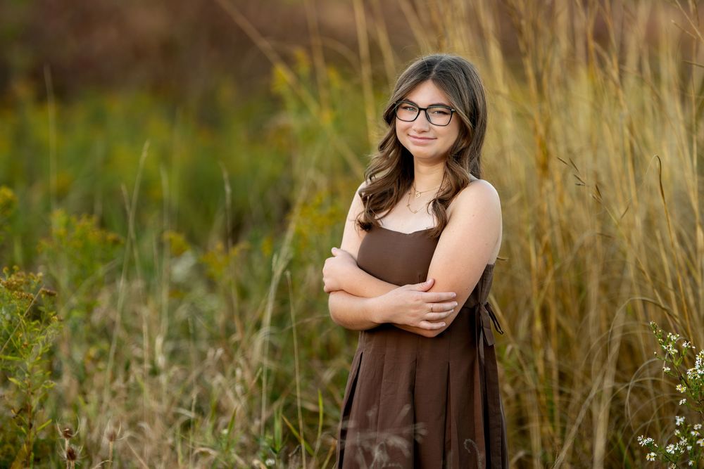 Teen girl in brown dress with arms crossed smiling in a field of tall grass.