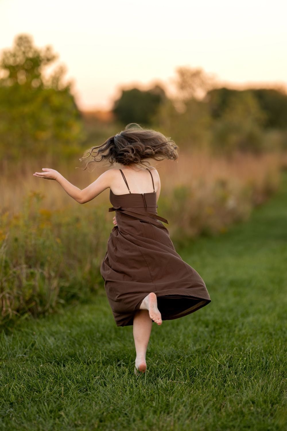 Young woman in brown dress dancing barefoot on grass path at golden sunset.
