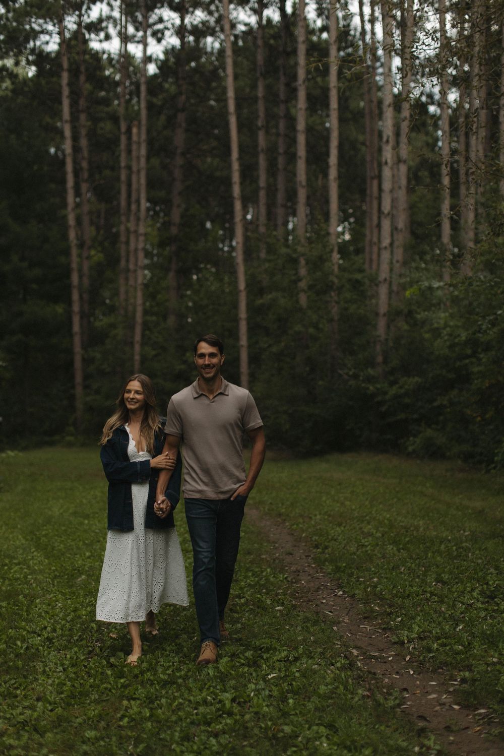 A couple walks along a forest trail surrounded by tall trees.