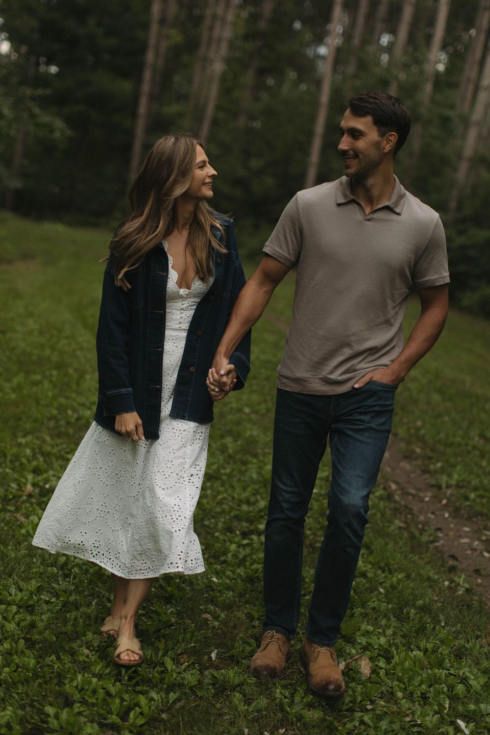 A couple strolls through a grassy clearing in a pine forest.