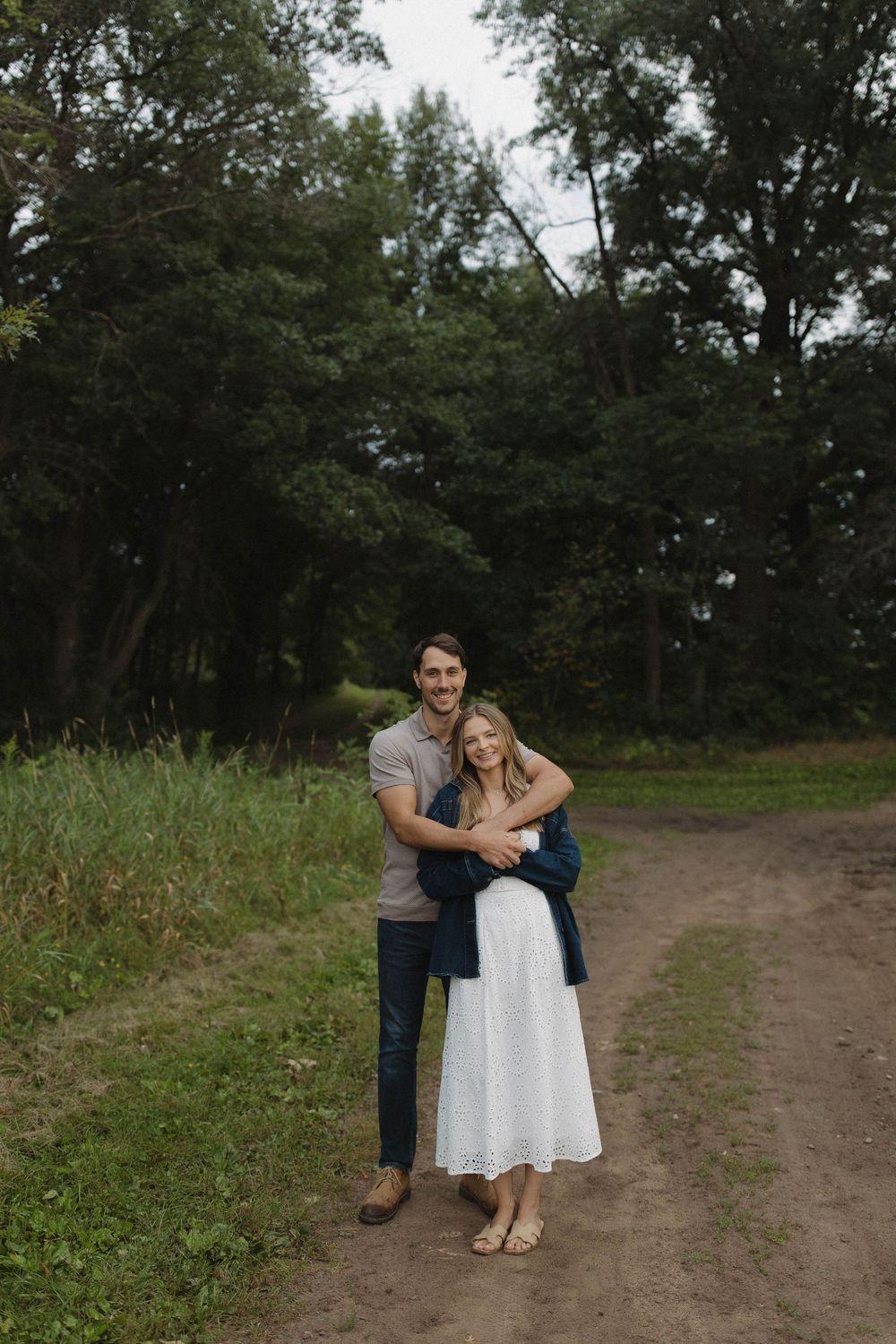 Romantic couple embracing on a dirt path surrounded by green trees.