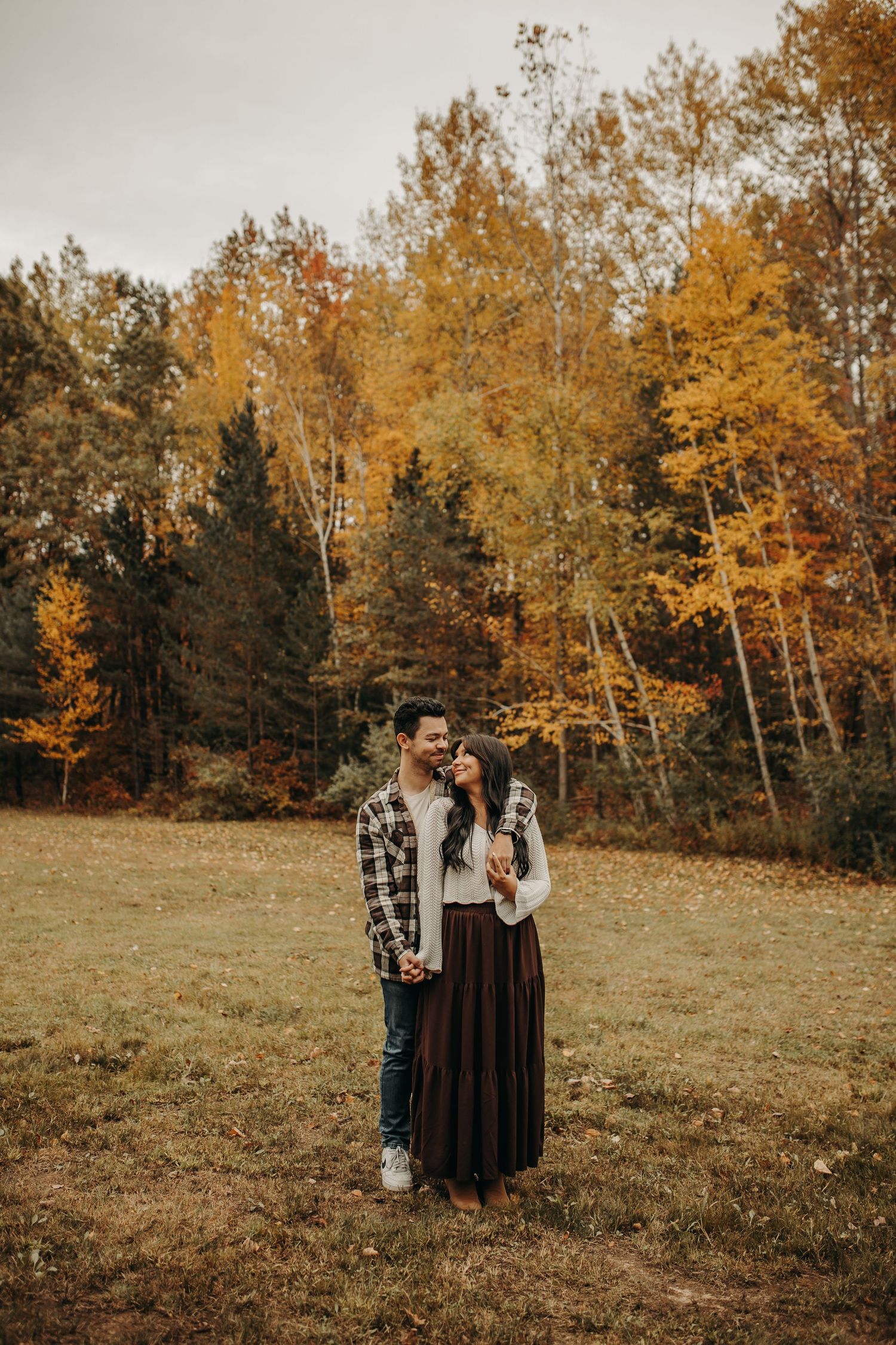 A couple stands together in a golden autumn field surrounded by trees with warm fall foliage at sunset. Creative Art By Bree LLC
