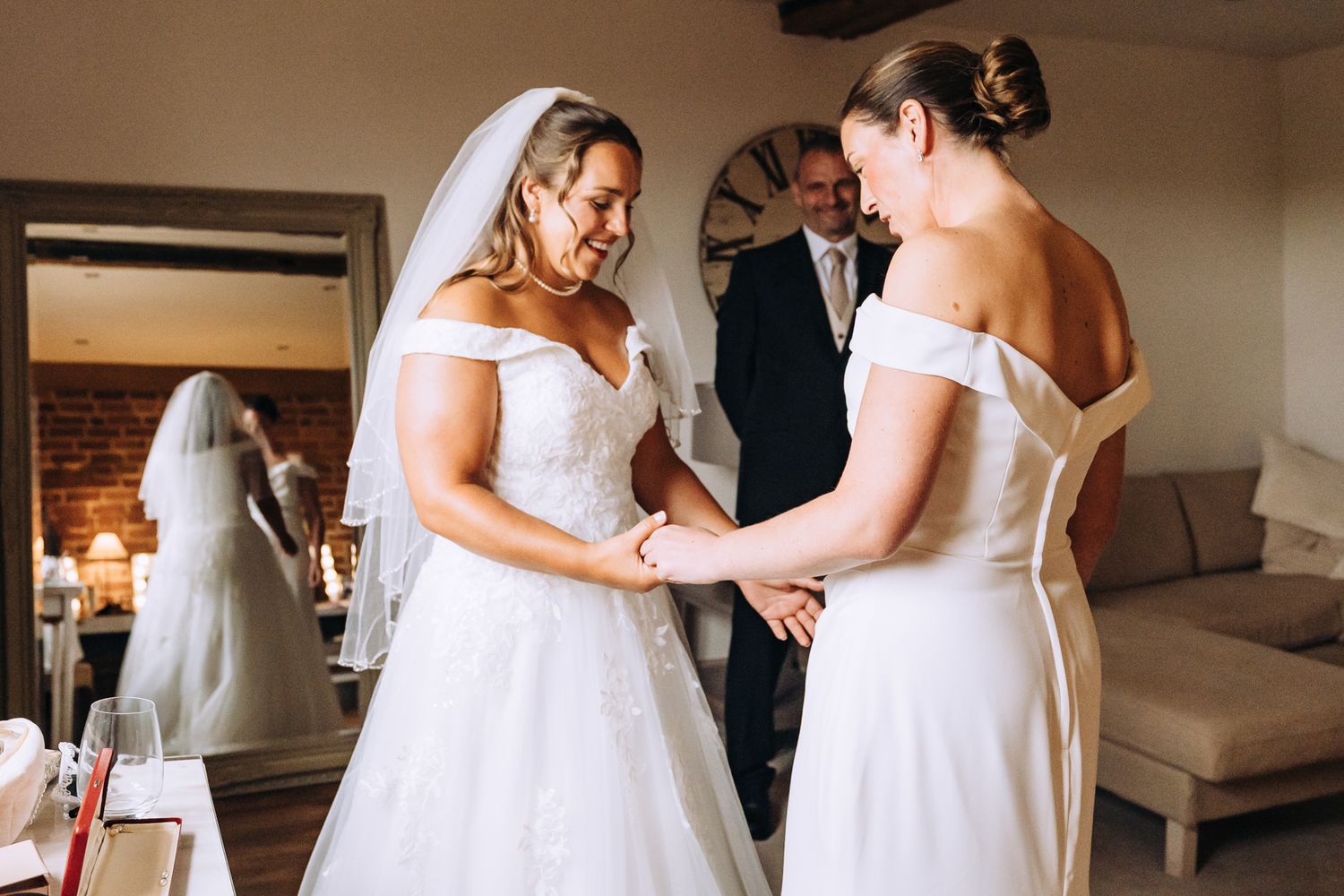 Two people in white dresses share an intimate moment during wedding preparations.