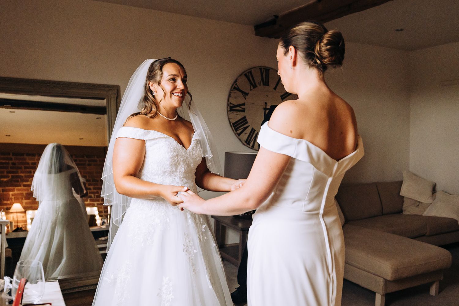 Two people in white dresses share a tender moment during wedding preparations.